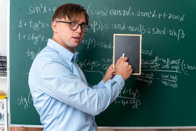 Joven profesor con gafas sosteniendo una pequeña pizarra y tiza sorprendido de pie cerca de la pizarra con fórmulas matemáticas en el aula