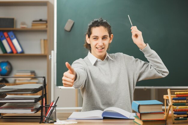 un joven profesor sonriente apunta a la pizarra con un puntero sentado en el escritorio mostrando los pulgares hacia arriba con herramientas escolares en el aula