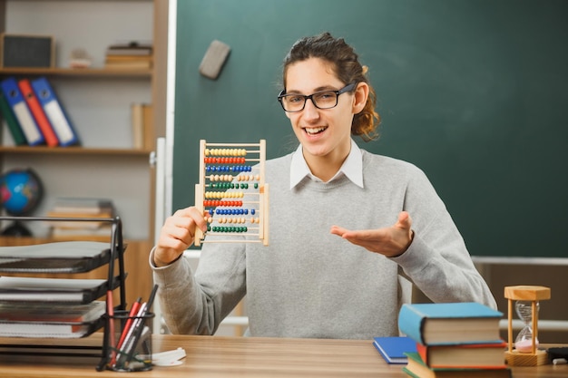 un joven profesor sonriente con gafas sentado en un escritorio sosteniendo y apuntando a un ábaco con herramientas escolares en el aula