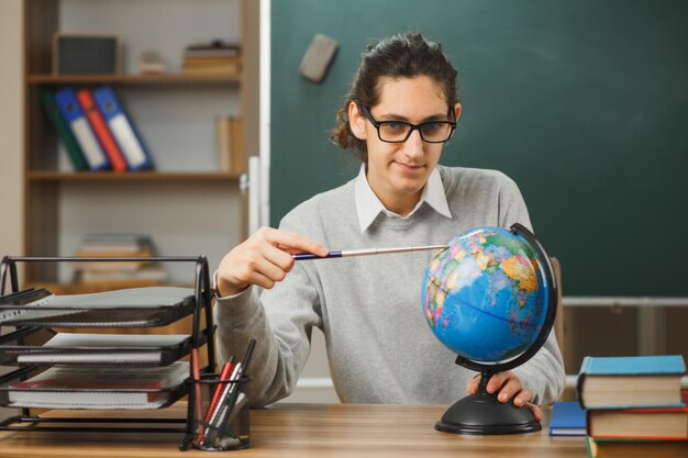 un joven profesor sonriente con gafas sentado en un escritorio sosteniendo un puntero con un globo terráqueo con herramientas escolares en el aula