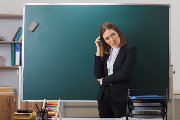 joven profesora con anteojos parada cerca de la pizarra en el aula explicando la lección luciendo confundida rascándose la cabeza tratando de recordar algo importante