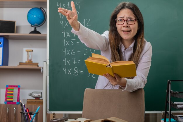 Joven profesora con gafas leyendo un libro explicando la lección sosteniendo la mano estar confundido y disgustado de pie en el escritorio de la escuela frente a la pizarra en el aula