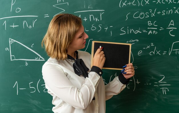 Joven profesora insatisfecha de pie delante de la pizarra sosteniendo y mirando la mini pizarra con varados para el tablero en el aula