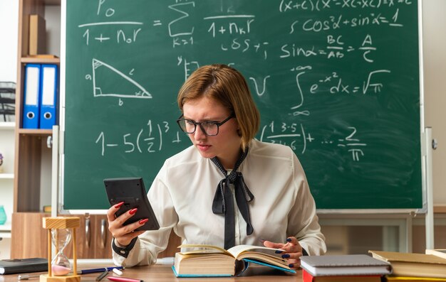 Joven profesora insatisfecha se sienta a la mesa con útiles escolares sosteniendo y mirando la calculadora en el aula