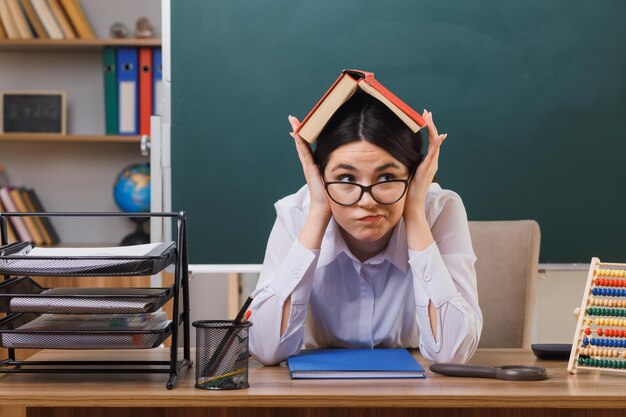 una joven profesora pensante sosteniendo un libro en la cabeza sentada en un escritorio con herramientas escolares en el aula