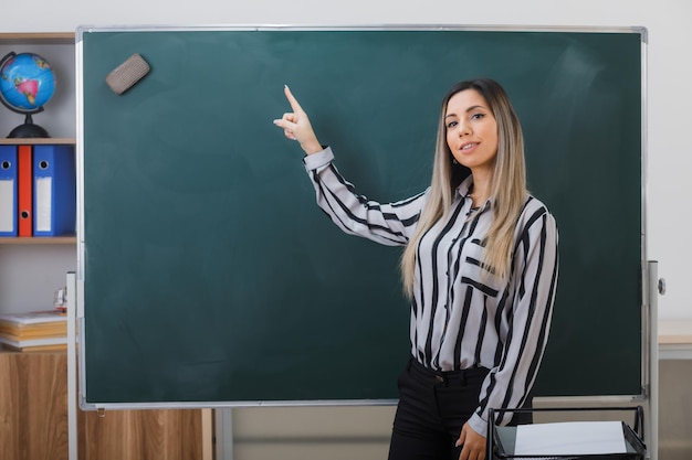 Foto gratuita joven profesora de pie cerca de la pizarra en el aula explicando la lección apuntando a la pizarra con el dedo índice mirando confiada