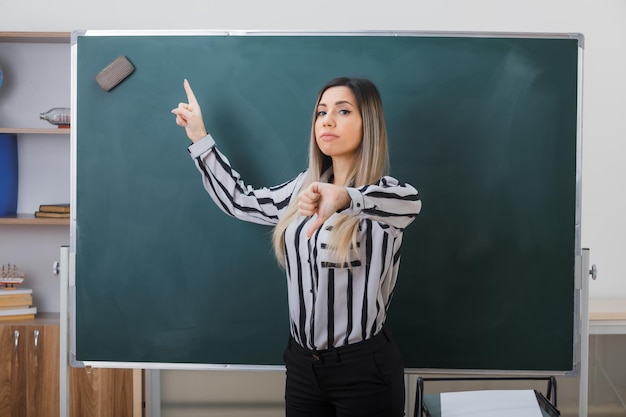 joven profesora de pie cerca de la pizarra en el aula explicando la lección apuntando a la pizarra con el dedo índice mostrando el pulgar hacia abajo con aspecto insatisfecho