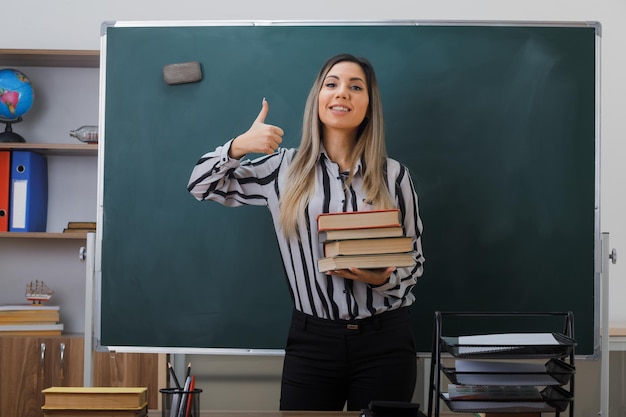 joven profesora de pie cerca de la pizarra en el aula explicando la lección sosteniendo una pila de libros mostrando el pulgar hacia arriba mirando a la cámara feliz y complacida sonriendo