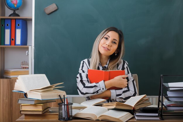 joven profesora sentada en el escritorio de la escuela frente a la pizarra en el aula sosteniendo un libro mirando a la cámara sonriendo amable, feliz y positiva