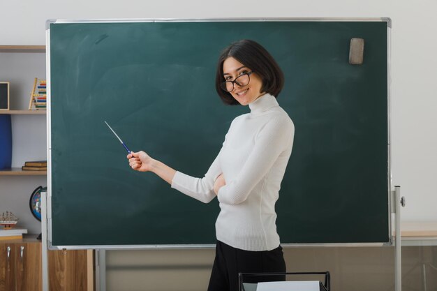 una joven profesora sonriente con anteojos parada frente a la pizarra y apuntando a la pizarra con un puntero en el aula