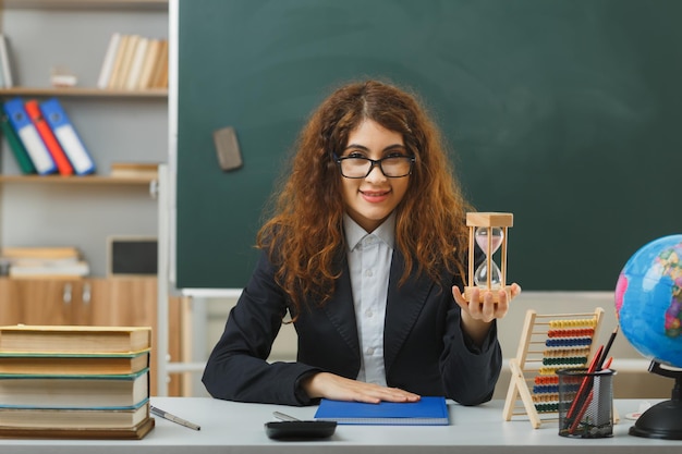 una joven profesora sonriente con gafas sosteniendo un reloj de arena sentada en un escritorio con herramientas escolares en el aula