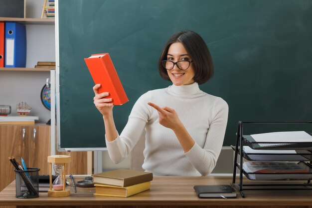 joven profesora sonriente con gafas sosteniendo y señalando el libro sentado en el escritorio con herramientas escolares en el aula