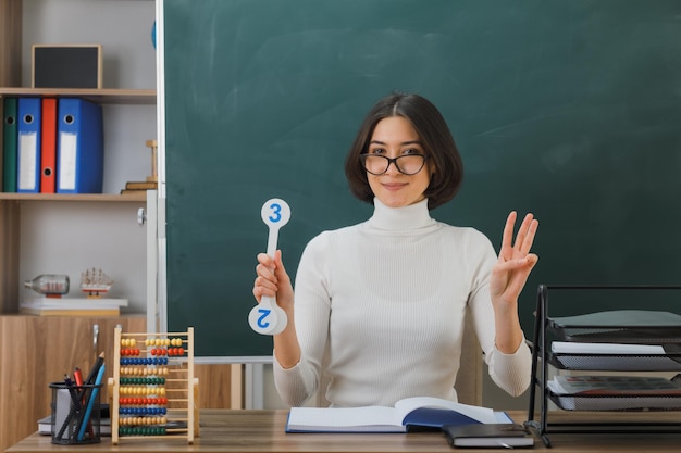 joven profesora sonriente con gafas sosteniendo un ventilador sentado en el escritorio con herramientas escolares en el aula