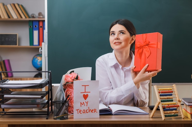 una joven profesora sonriente sosteniendo un regalo sentada en un escritorio con herramientas escolares en el aula