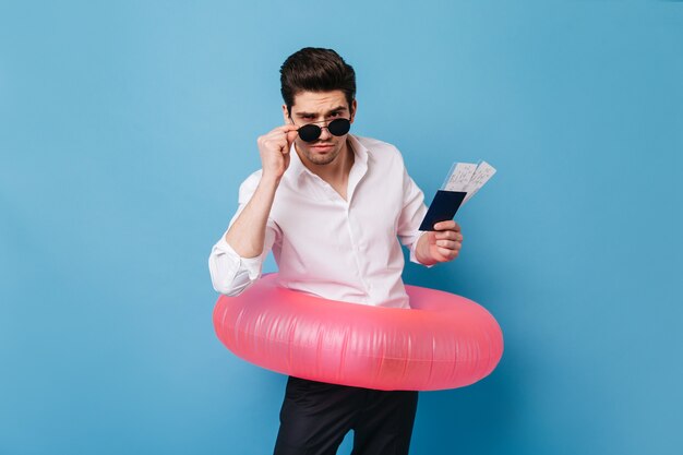 Joven se quita las gafas de sol. Guy tiene documentos y boletos. Morena con camisa blanca mira a cámara y posa con círculo inflable.