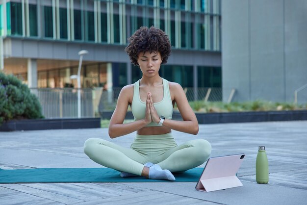 Joven relajada hace yoga en la colchoneta se sienta en posición de loto mantiene las palmas juntas los ojos cerrados respira profundamente mira el video tutorial