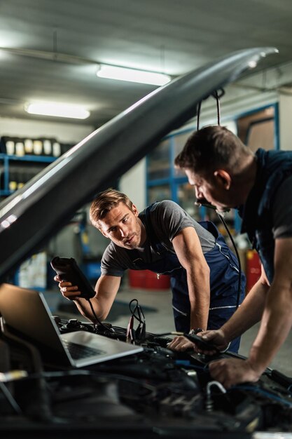 Joven reparador de automóviles hablando con su compañero de trabajo mientras revisa el rendimiento del motor con herramientas de diagnóstico en el taller de reparación de automóviles