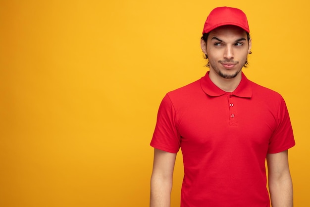 un joven repartidor astuto con uniforme y gorra mirando al costado con algunos planes malvados aislados en un fondo amarillo con espacio para copiar