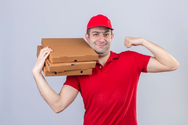 Joven repartidor en camisa polo roja y gorra sosteniendo cajas de pizza levantando el puño sonriendo seguro ganador concepto sobre pared blanca aislada