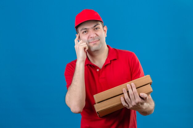 Joven repartidor en camisa polo roja y gorra sosteniendo cajas de pizza de pie con la cara ceñuda apuntando a la cabeza con dolor de cabeza sobre la pared azul aislada