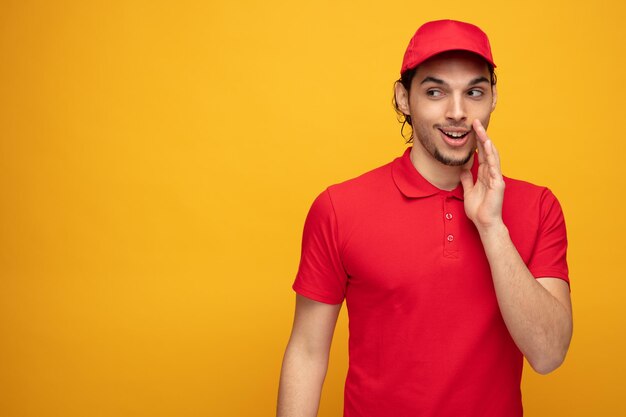 joven repartidor complacido con uniforme y gorra manteniendo la mano cerca de la boca mirando al costado susurrando aislado en fondo amarillo con espacio para copiar