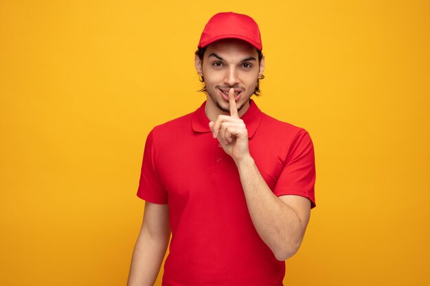 joven repartidor complacido con uniforme y gorra mirando a la cámara mostrando un gesto de silencio aislado en un fondo amarillo