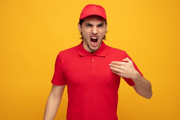 joven repartidor enojado con uniforme y gorra mirando a la cámara apuntándose a sí mismo con la mano gritando aislado en el fondo amarillo