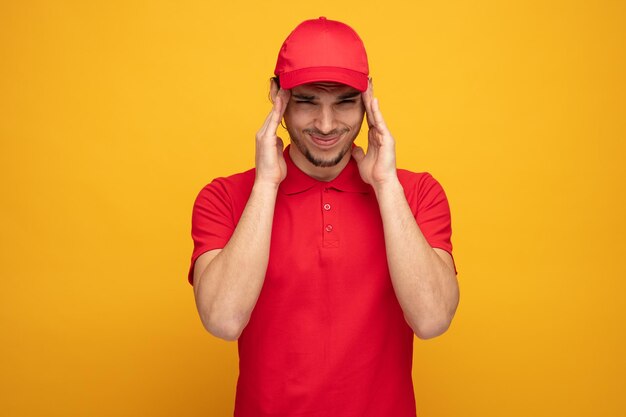 joven repartidor frunciendo el ceño con uniforme y gorra manteniendo las manos en la cabeza mirando a la cámara con dolor de cabeza aislado en el fondo amarillo