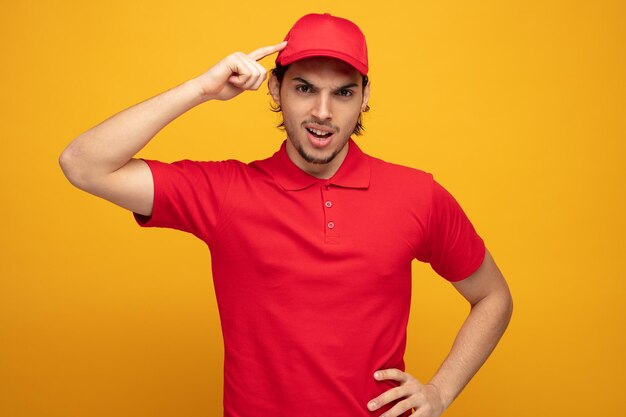 joven repartidor frunciendo el ceño con uniforme y gorra mirando a la cámara manteniendo la mano en la cintura mostrando si eres un gesto loco aislado en el fondo amarillo