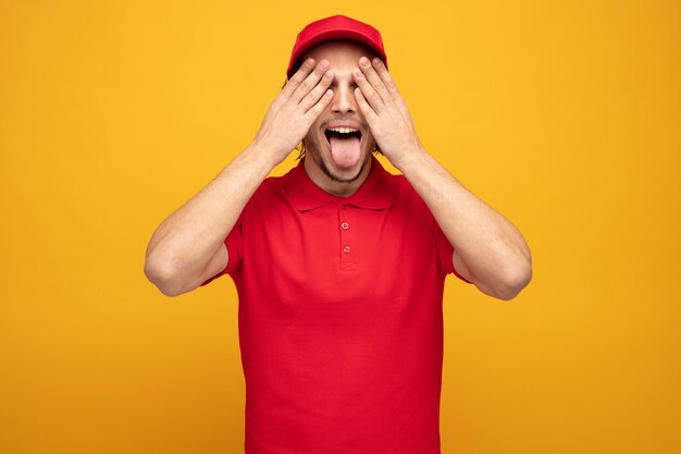 joven repartidor juguetón con uniforme y gorra cerrando los ojos con las manos mostrando la lengua aislada en el fondo amarillo