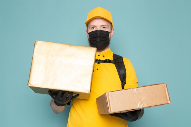 joven repartidor en polo amarillo gorra amarilla jeans blancos mochila y máscara estéril negra sosteniendo una caja en azul