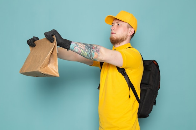 joven repartidor en polo amarillo gorra amarilla jeans blancos mochila sosteniendo una caja en azul