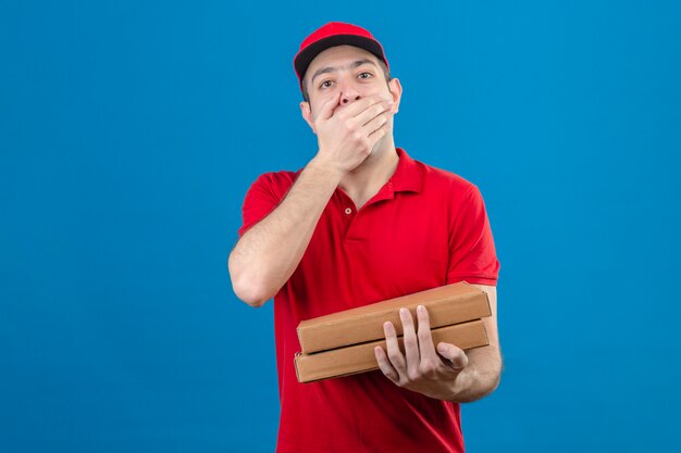 Joven repartidor en polo rojo y gorra con cajas de pizza cubriendo la boca con la mano sorprendida de pie sobre la pared azul aislada