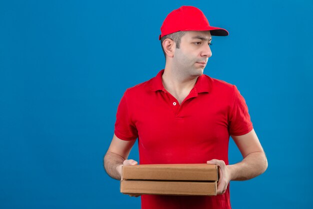 Joven repartidor en polo rojo y gorra con cajas de pizza mirando hacia el lado con cara seria de pie sobre la pared azul aislada