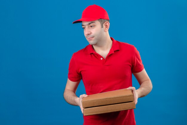 Joven repartidor en polo rojo y gorra con cajas de pizza mirando hacia el lado con cara seria sobre pared azul aislado