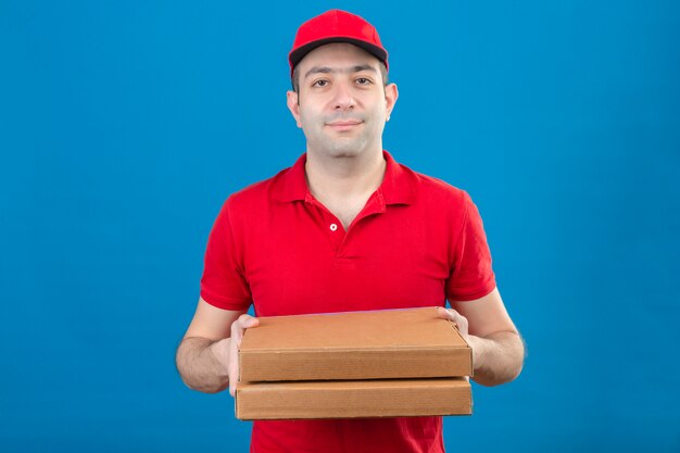 Joven repartidor en polo rojo y gorra con cajas de pizza sonriendo amable de pie sobre la pared azul aislado