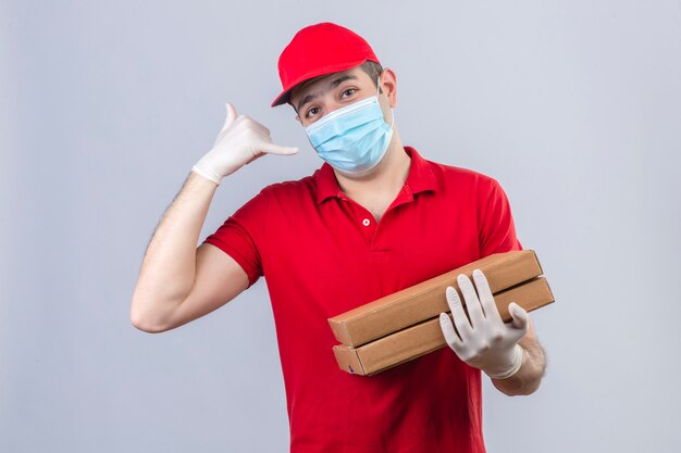 Joven repartidor en polo rojo y gorra en máscara médica sosteniendo cajas de pizza haciendo llamarme gesto mirando confiado sobre pared blanca aislada