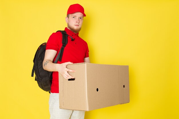 joven repartidor en polo rojo gorra roja jeans blancos sosteniendo una caja en amarillo