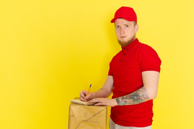 joven repartidor en polo rojo gorra roja jeans blancos sosteniendo una caja y escribiendo en amarillo