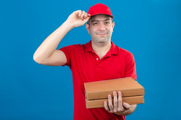 Joven repartidor en polo rojo y gorra sosteniendo cajas de pizza haciendo gesto de saludo tocando su gorra sonrisa confiada en la cara sobre la pared azul aislada