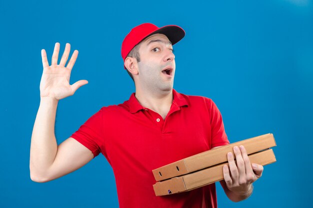 Joven repartidor en polo rojo y gorra sosteniendo cajas de pizza saludando con la mano mirando sorprendido sobre la pared azul aislada