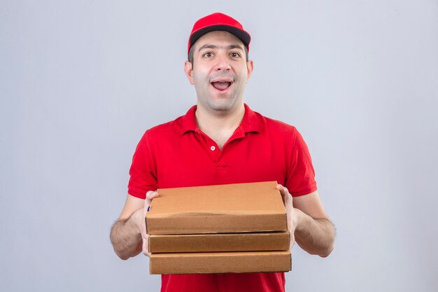 Joven repartidor en polo rojo y gorra sosteniendo cajas de pizza sonriendo alegremente de pie sobre la pared blanca aislada