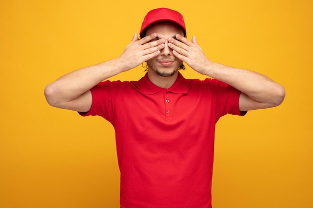 un joven repartidor serio con uniforme y gorra cerrando los ojos con las manos aisladas en el fondo amarillo