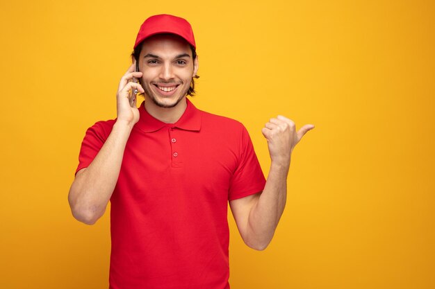 un joven repartidor sonriente con uniforme y gorra mirando a la cámara apuntando hacia un lado mientras habla por teléfono aislado de fondo amarillo