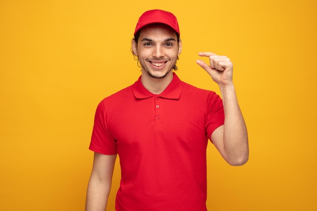 Foto gratuita un joven repartidor sonriente con uniforme y gorra mirando a la cámara mostrando un pequeño gesto aislado de fondo amarillo