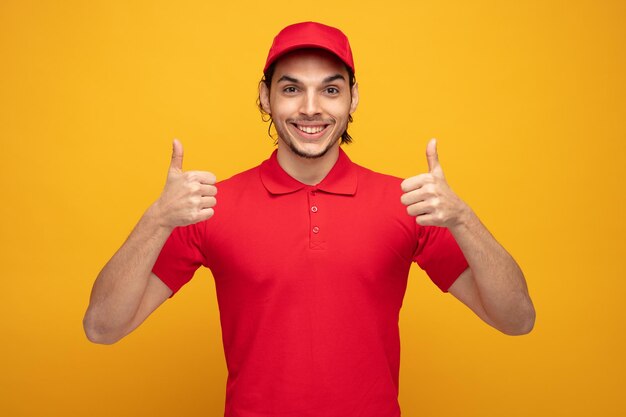 un joven repartidor sonriente con uniforme y gorra mirando a la cámara mostrando los pulgares hacia arriba aislado en el fondo amarillo