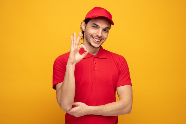 un joven repartidor sonriente con uniforme y gorra mirando a la cámara mostrando el signo de ok mientras mantiene la mano bajo el codo aislada en el fondo amarillo