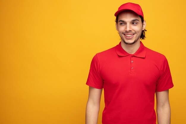 un joven repartidor sonriente con uniforme y gorra mirando a un lado aislado en un fondo amarillo con espacio para copiar