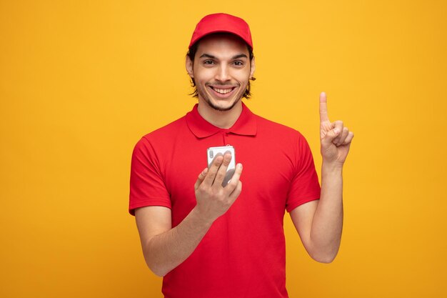 un joven repartidor sonriente con uniforme y gorra sosteniendo un teléfono móvil mirando a la cámara apuntando hacia arriba aislado en un fondo amarillo