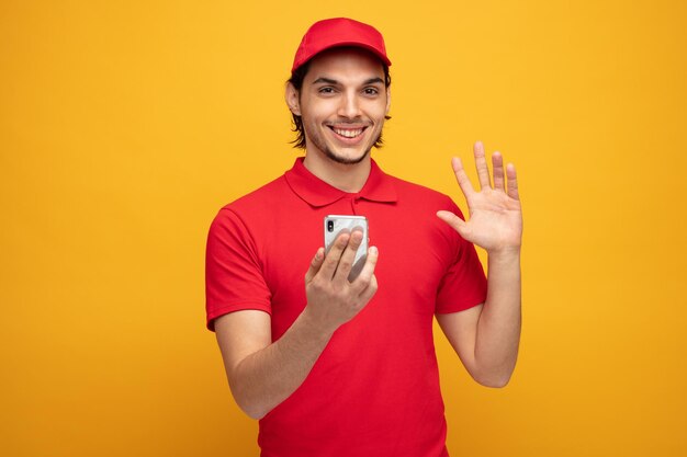 un joven repartidor sonriente con uniforme y gorra sosteniendo un teléfono móvil mirando a la cámara saludando aislado de fondo amarillo